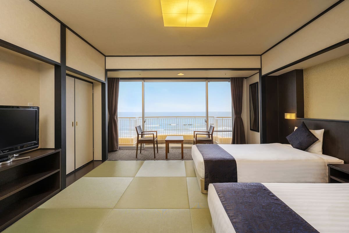 A hotel room with two beds, a TV, a balcony with a view of the sea, and a table with chairs.