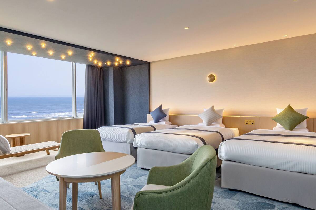 A hotel room with two beds, a small table, and a chair. There is a view of the ocean through large windows.