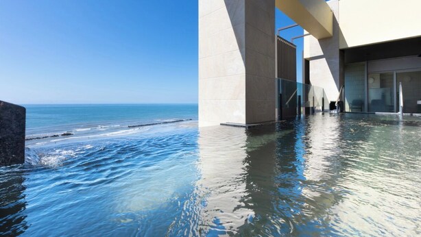 Una piscina infinita con vistas al océano, un edificio moderno con amplios ventanales y un cielo azul despejado.