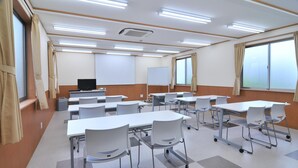 Une salle de conférence avec des tables et des chaises blanches, un tableau blanc, un projecteur et un téléviseur.