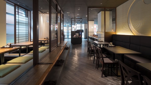 A modern restaurant interior with wooden tables and benches, a central counter, and large windows allowing natural light. - Richmond Hotel Kumamoto Shinshigai (Kumamoto)