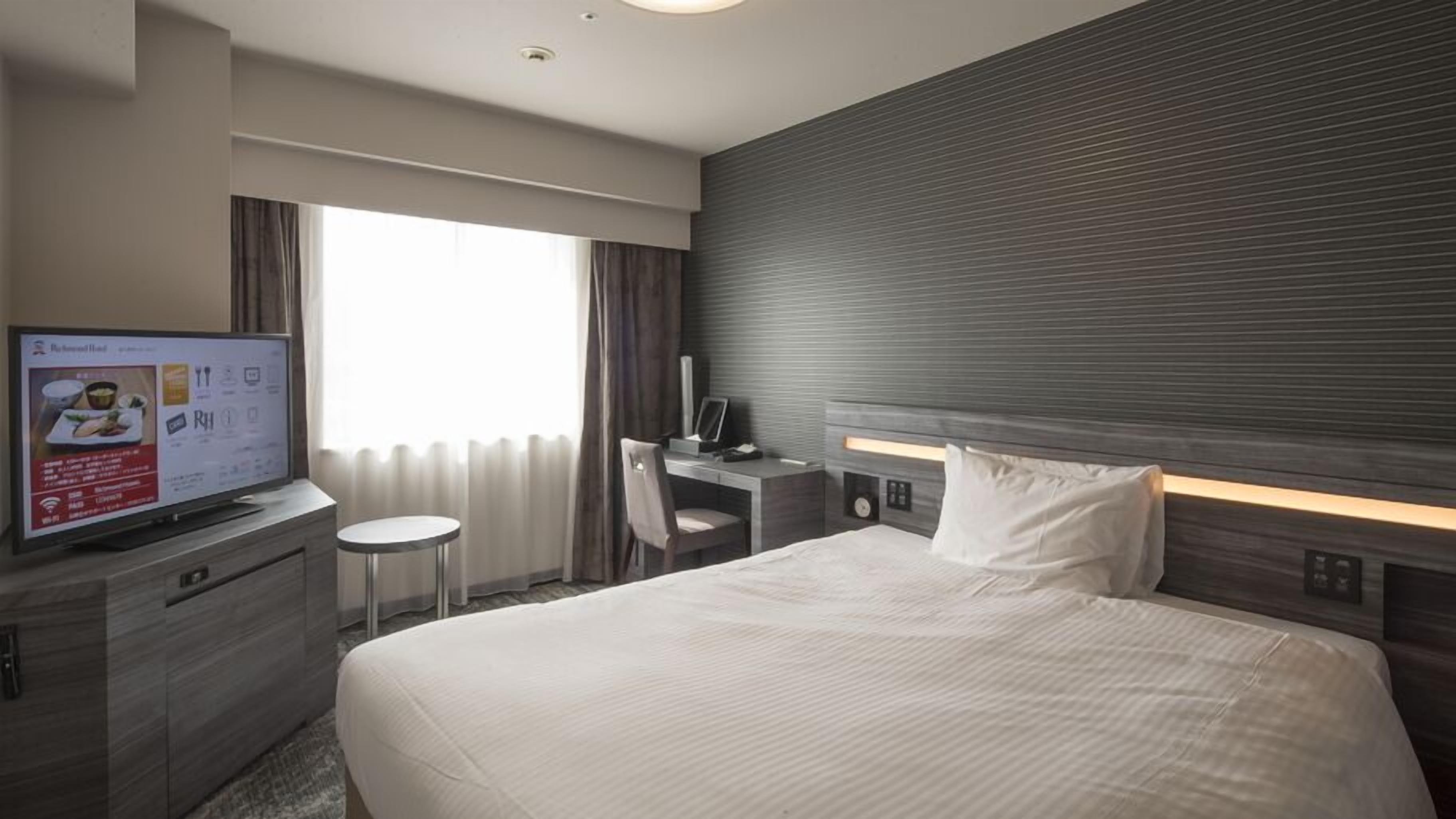 Habitación de hotel con cama, escritorio, silla, televisión y ventana con cortinas.