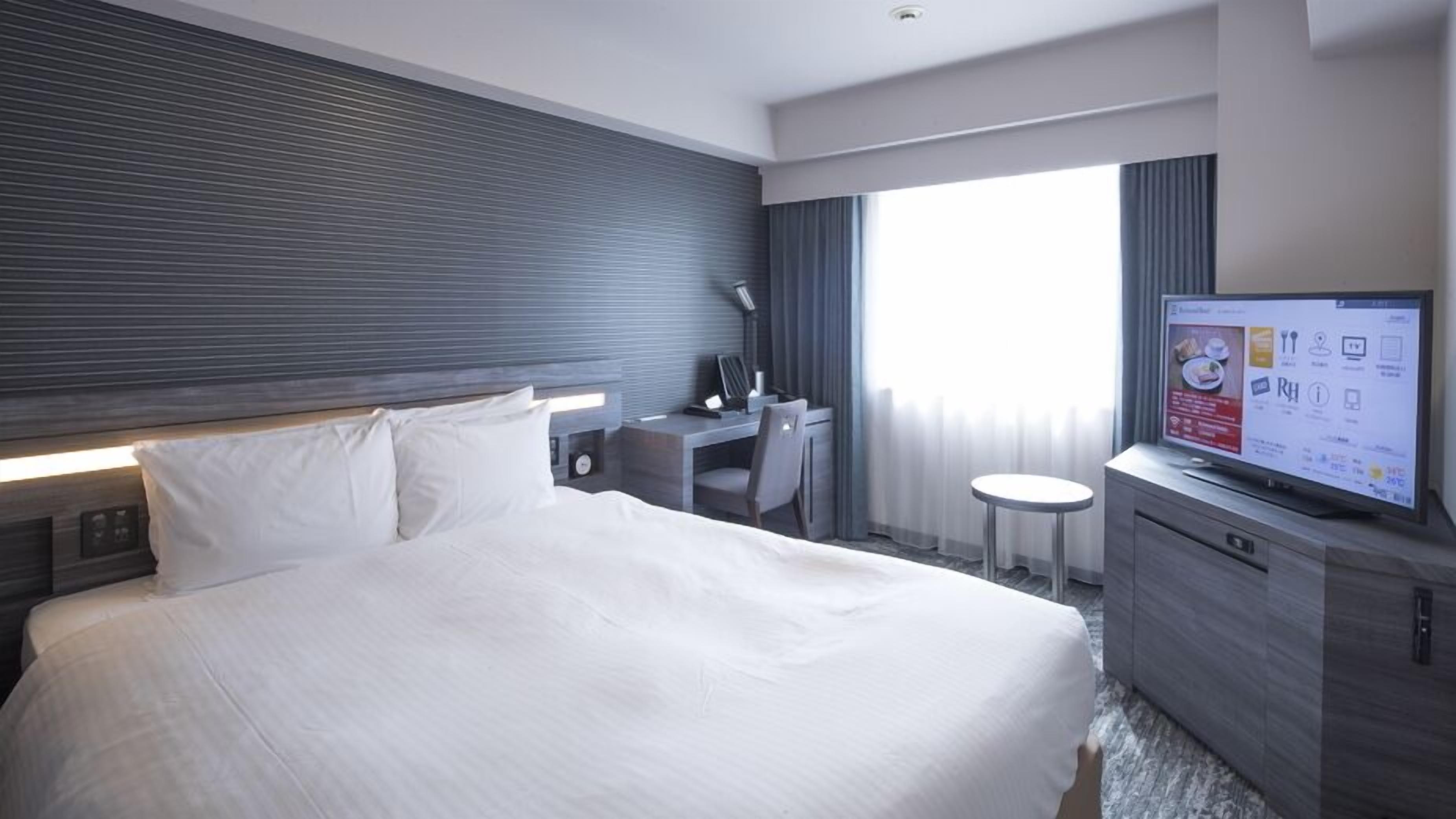Habitación de hotel con cama, escritorio, silla, televisor y ventana con cortinas.