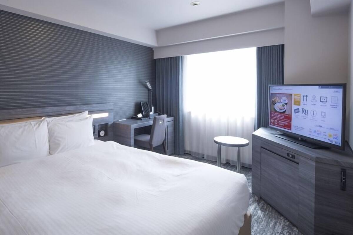 A hotel room with a bed, a desk, a chair, a TV, and a window with curtains.