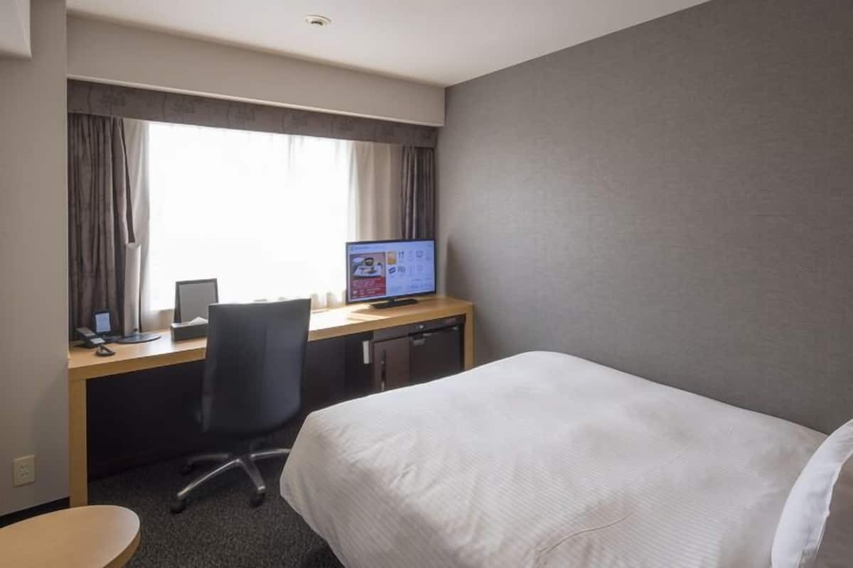 A hotel room with a bed, a desk with a computer, a chair, and a window with curtains.