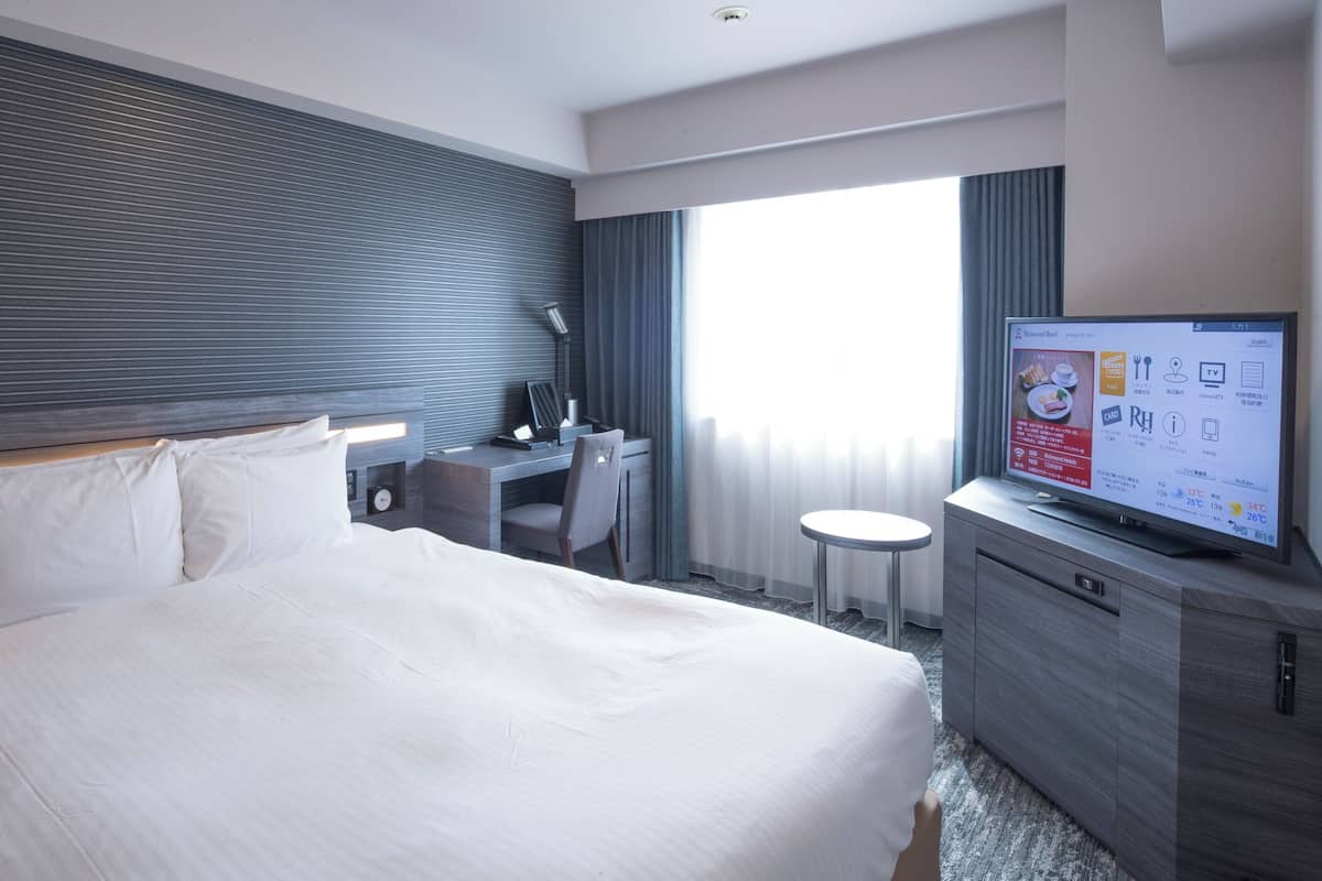 A modern hotel room with a large bed, a desk, a TV, and a view of the city.