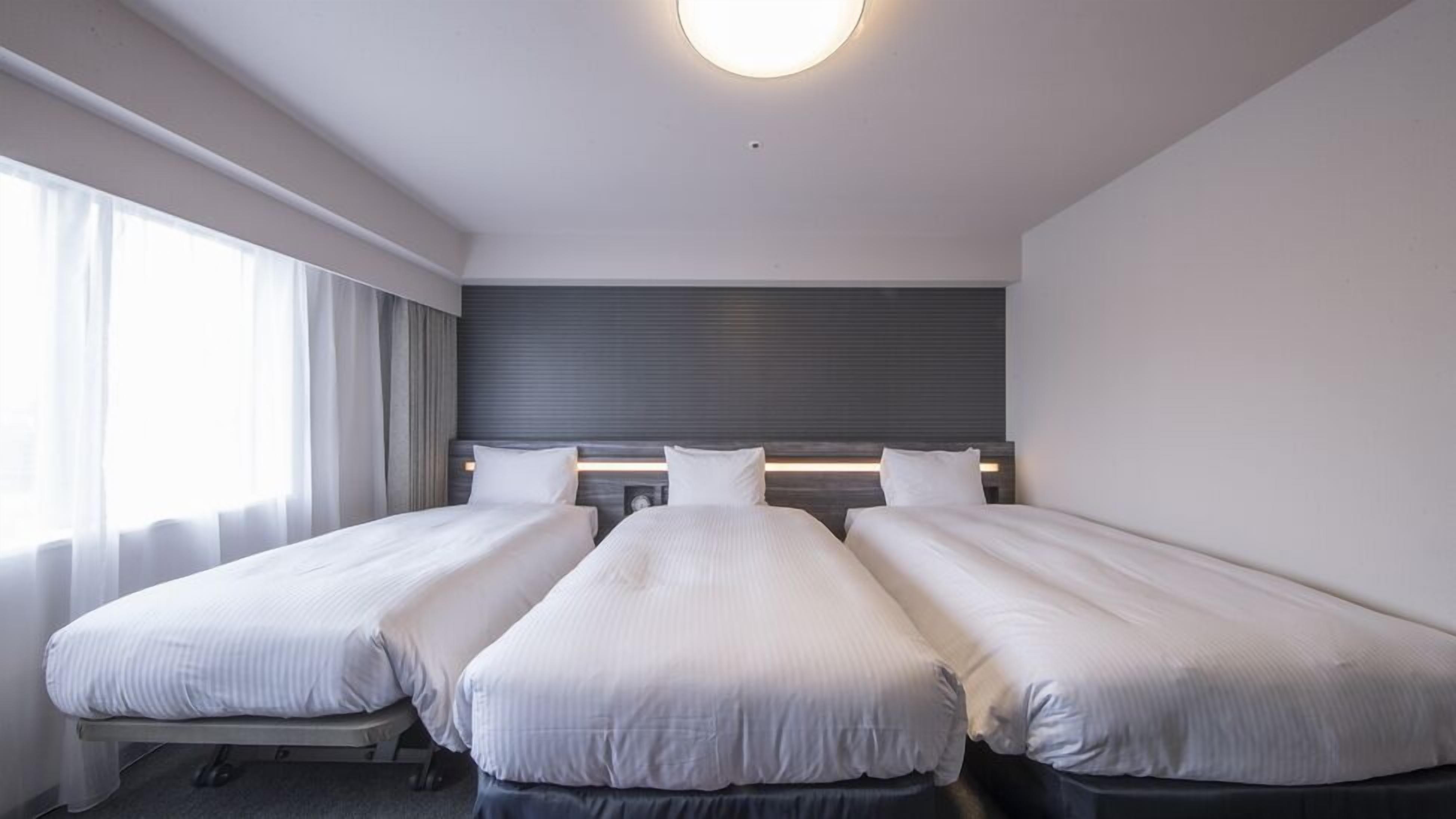 Habitación de hotel con tres camas individuales, un ventanal grande con cortinas, un cabecero moderno y una lámpara en el techo.