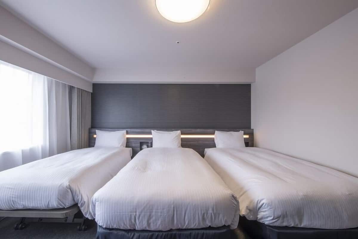 A hotel room with three single beds, a large window with curtains, a modern headboard, and a ceiling light.