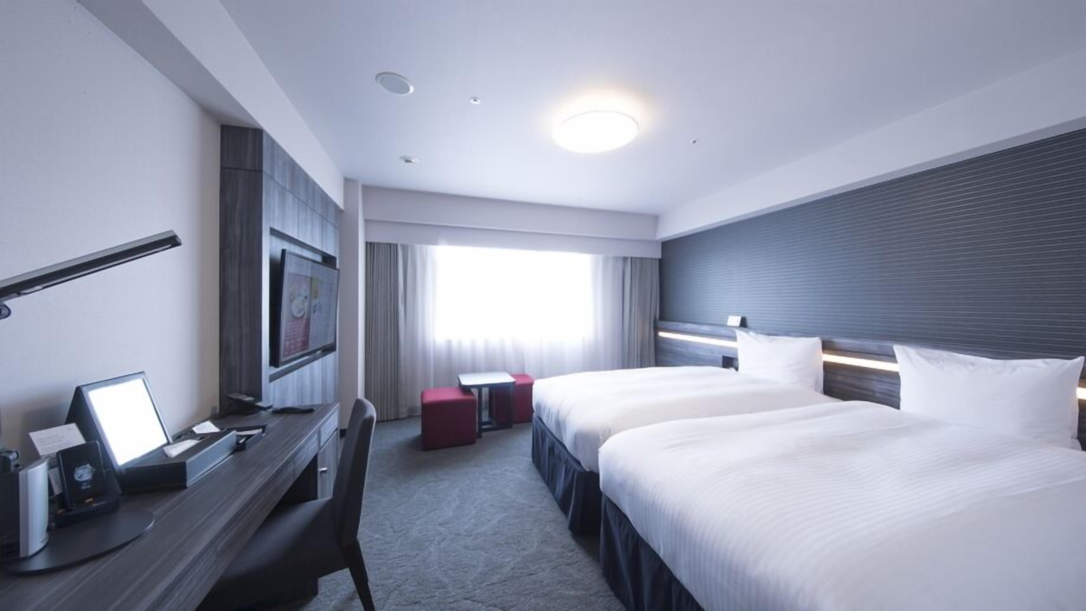 Habitación de hotel con dos camas, un escritorio con computadora, un televisor y una ventana con cortinas.