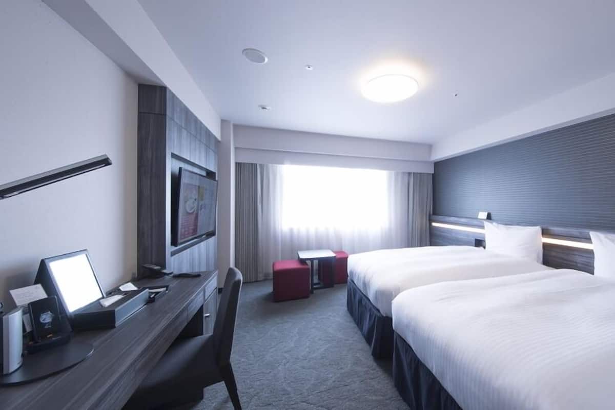 A hotel room with two beds, a desk with a computer, a TV, and a window with curtains.