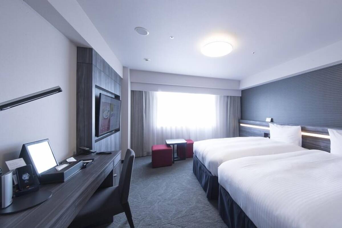 Habitación de hotel con dos camas, un escritorio con computadora, un televisor y una ventana con cortinas.