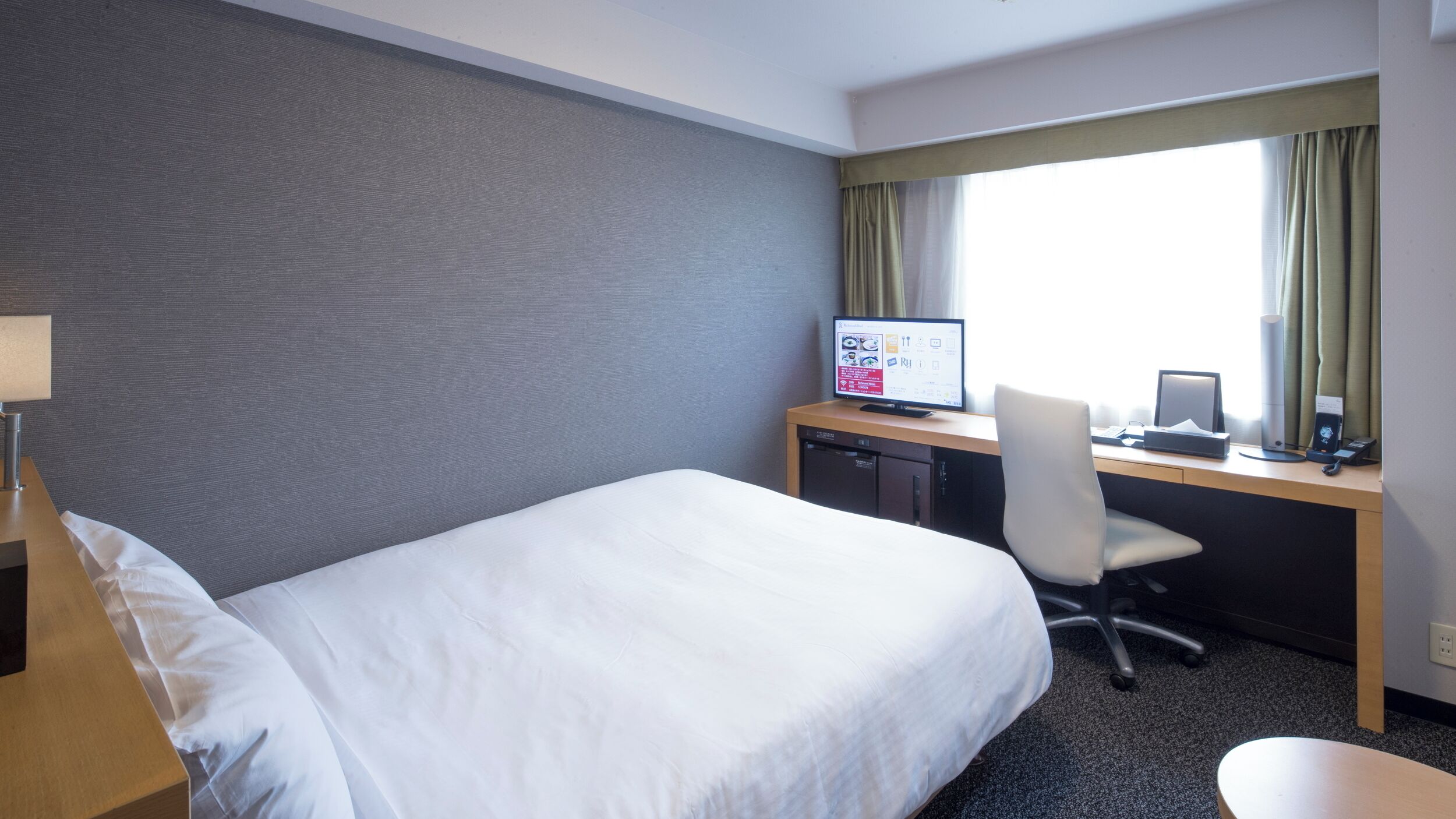 Habitación de hotel con cama, escritorio, computadora, silla y ventana con cortinas.