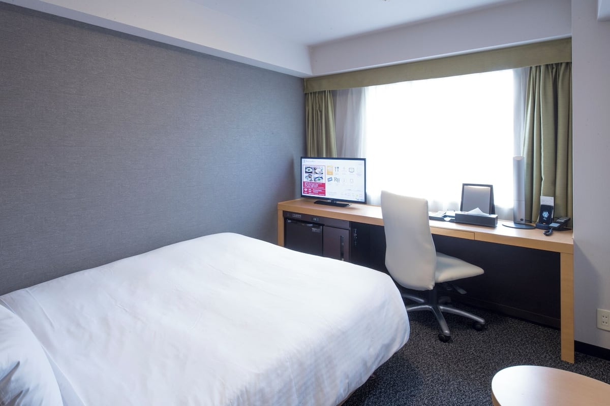 A hotel room with a bed, desk, computer, chair, and window with curtains.