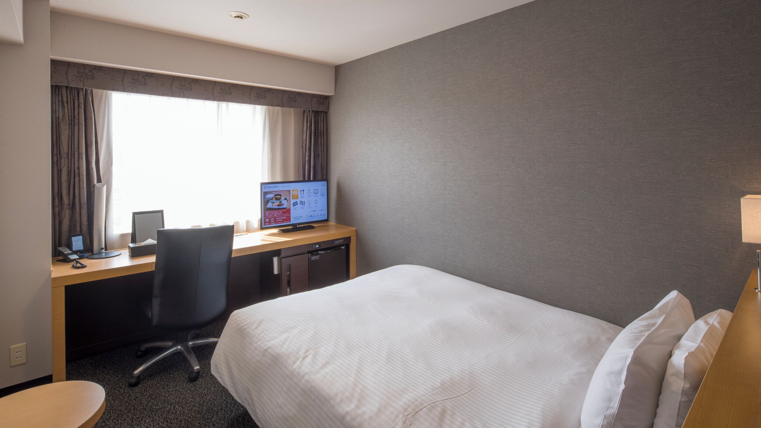 Habitación de hotel con cama, escritorio, computadora y ventana con cortinas.