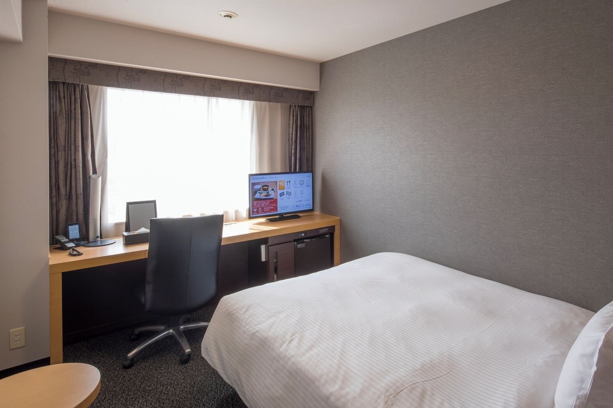 A hotel room with a bed, desk, computer, and window with curtains.