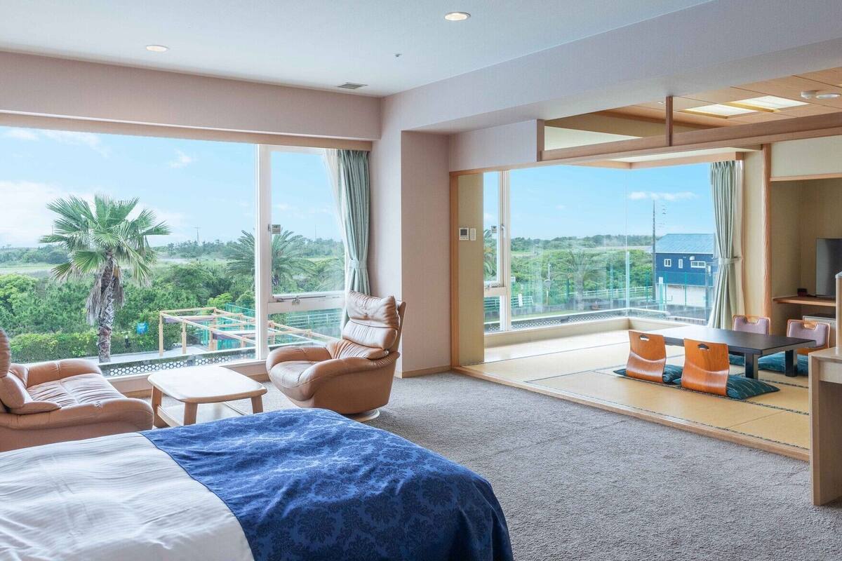 Une chambre d’hôtel comprenant un lit, deux chaises, une petite table, un bureau avec une chaise, et de grandes fenêtres donnant sur un paysage verdoyant et un immeuble.