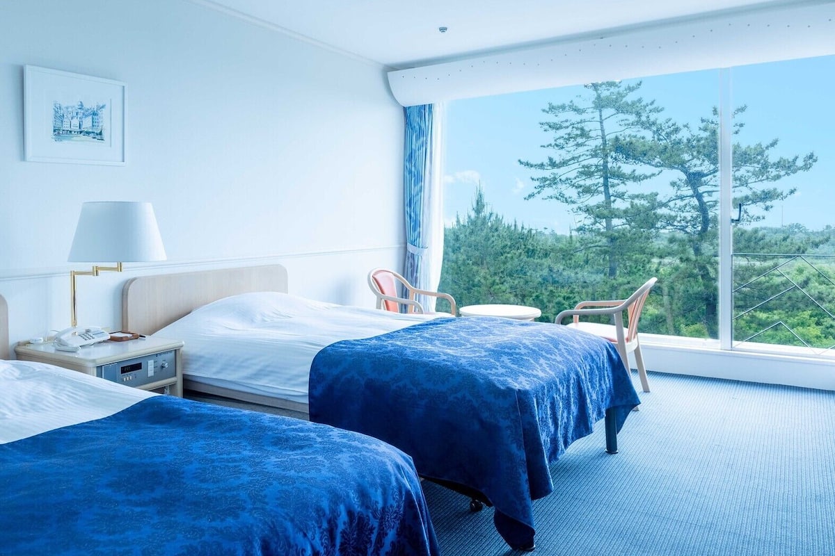 Une chambre d’hôtel avec deux lits, une table de chevet avec une lampe, une chaise et une vue sur les arbres par une grande fenêtre.