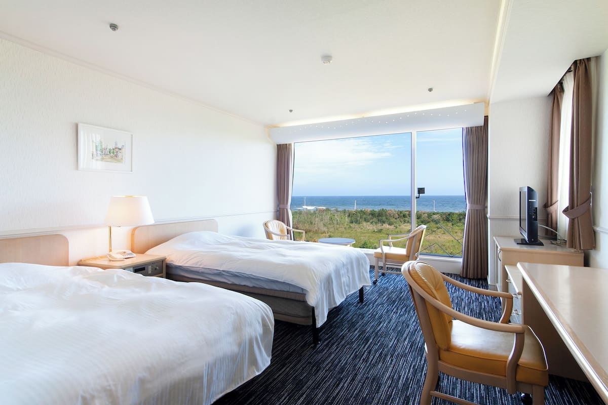 Une chambre d’hôtel avec deux lits, un bureau, une chaise, une télévision et une grande fenêtre donnant sur l’océan.