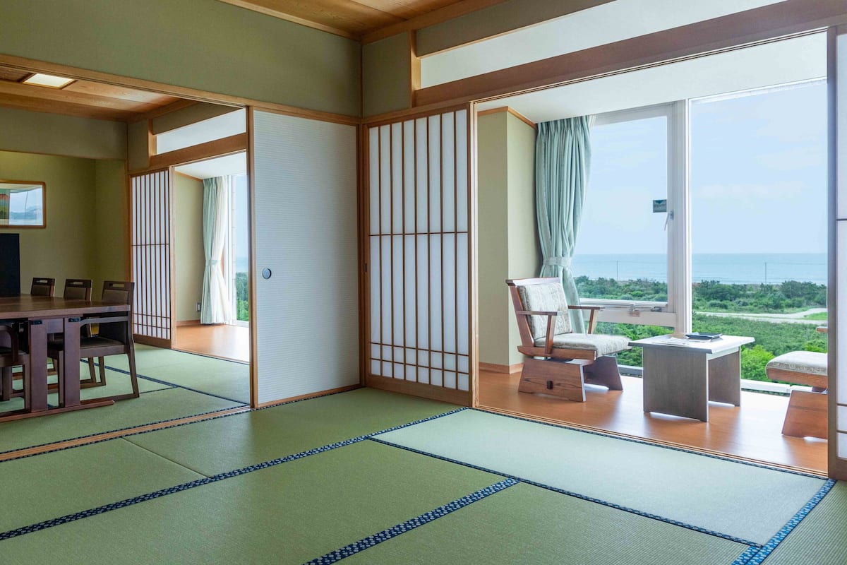 Une pièce de style japonais traditionnel, avec un sol recouvert de tatamis, un coin repas comprenant une table et des chaises, un téléviseur et une vue sur la mer grâce à de grandes fenêtres.
