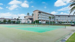 A tennis court with a net, surrounded by a building with multiple balconies and palm trees. - Hotel Ichinomiya Seaside Otsuka (Ichinomiya)