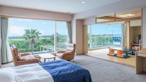 Habitación de hotel con cama, zona de estar con sillones, escritorio y amplios ventanales con vistas a zonas verdes y un edificio.
