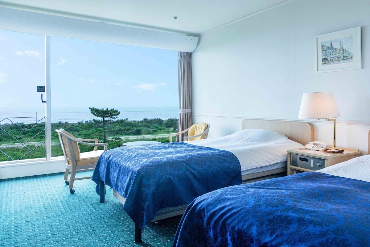 Une chambre d’hôtel avec deux lits, une chaise, une table de chevet avec une lampe et une vue sur la mer.