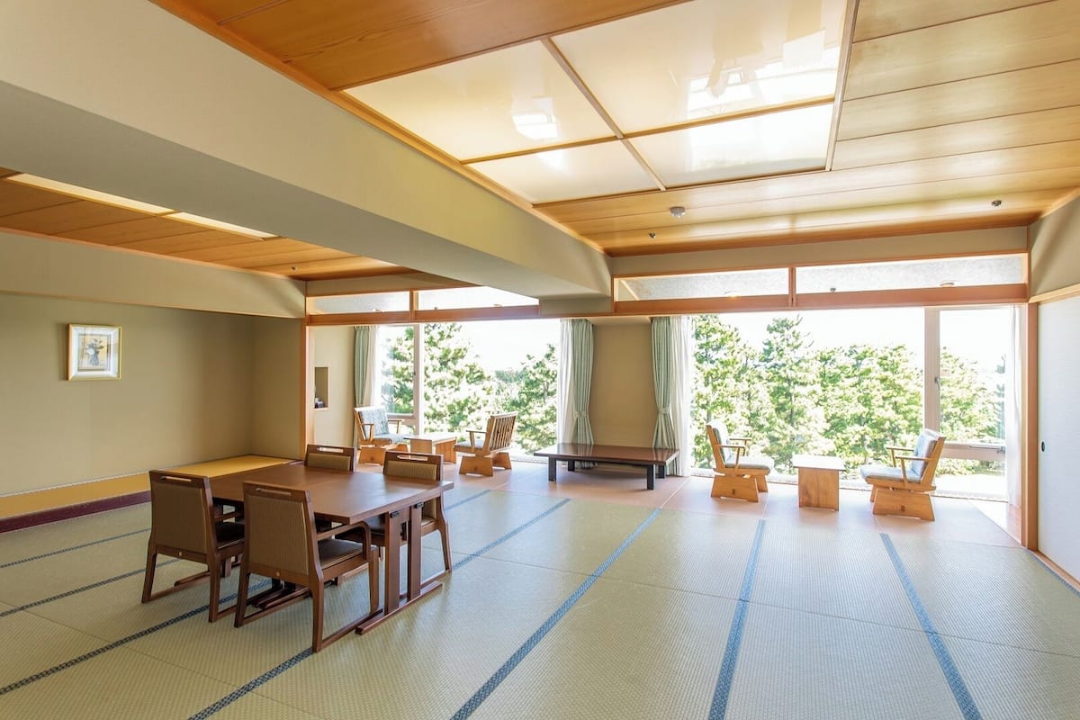 Une pièce avec un sol recouvert de tatamis, une petite table et des chaises, de grandes fenêtres et un plafond orné de poutres en bois.
