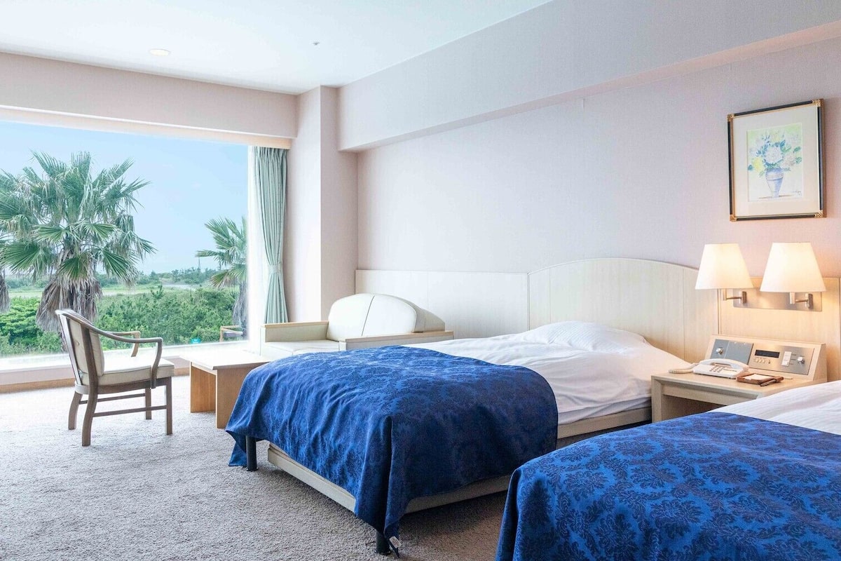 Une chambre d’hôtel avec deux lits, un bureau, une chaise, une table de chevet avec un téléphone et une fenêtre donnant sur des palmiers.