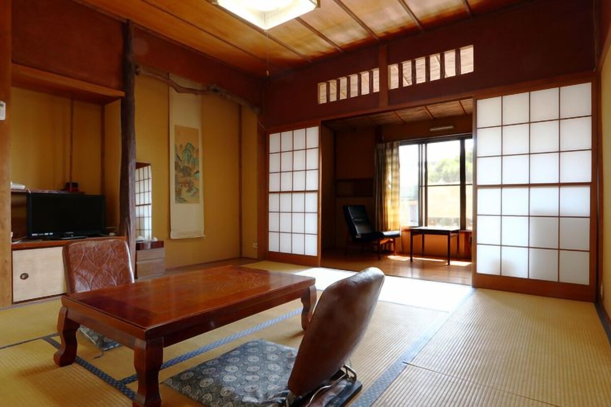 A traditional Japanese room with tatami flooring, a low wooden table, a cushioned chair, a television, and sliding shoji doors.