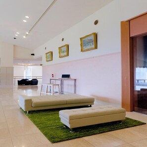 Una habitación de hotel moderna con sofá, reposapiés y una alfombra verde. Se observan cuadros en la pared y un escritorio con una computadora portátil.