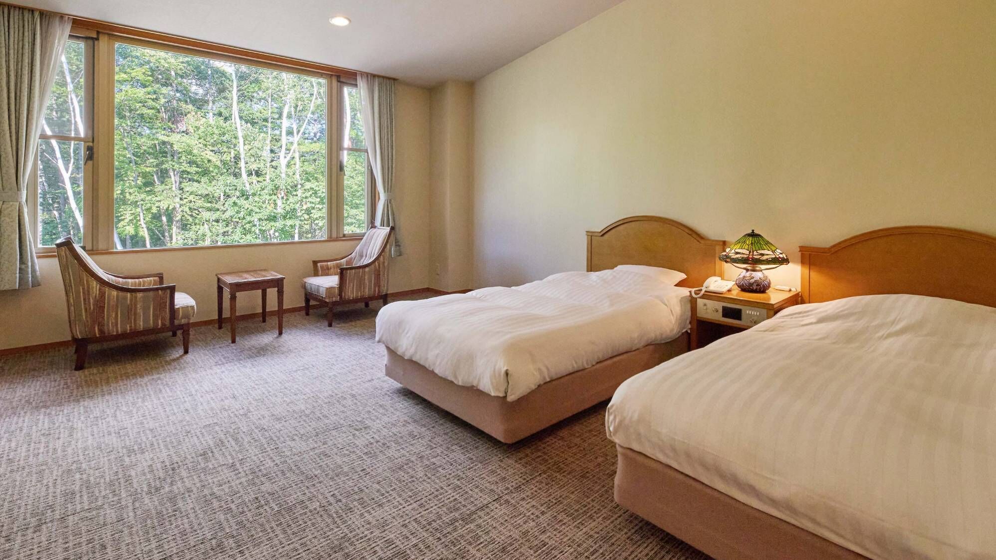 Een hotelkamer met twee bedden, een stoel, een tafeltje en een raam met uitzicht op bomen.