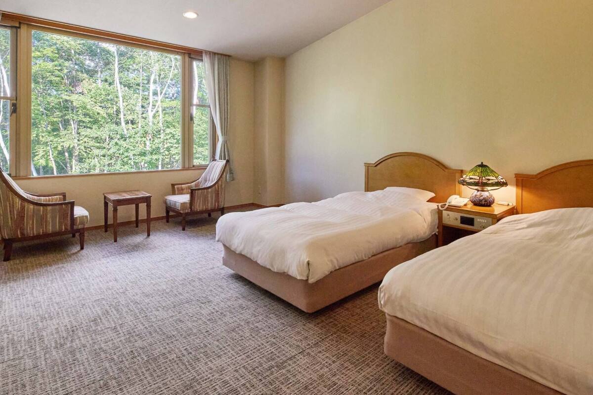 Ein Hotelzimmer mit zwei Betten, einem Sessel, einem kleinen Tisch und einem Fenster mit Blick auf Bäume.