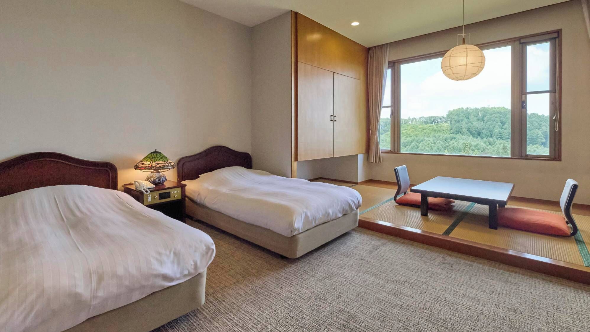 A hotel room with two beds, a small table, a chair, a lamp, and a view of greenery through the window.