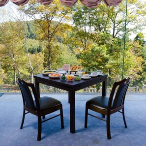 Un área de comedor al aire libre con una mesa dispuesta para dos personas, rodeada de sillas y vista a árboles y colinas al fondo.