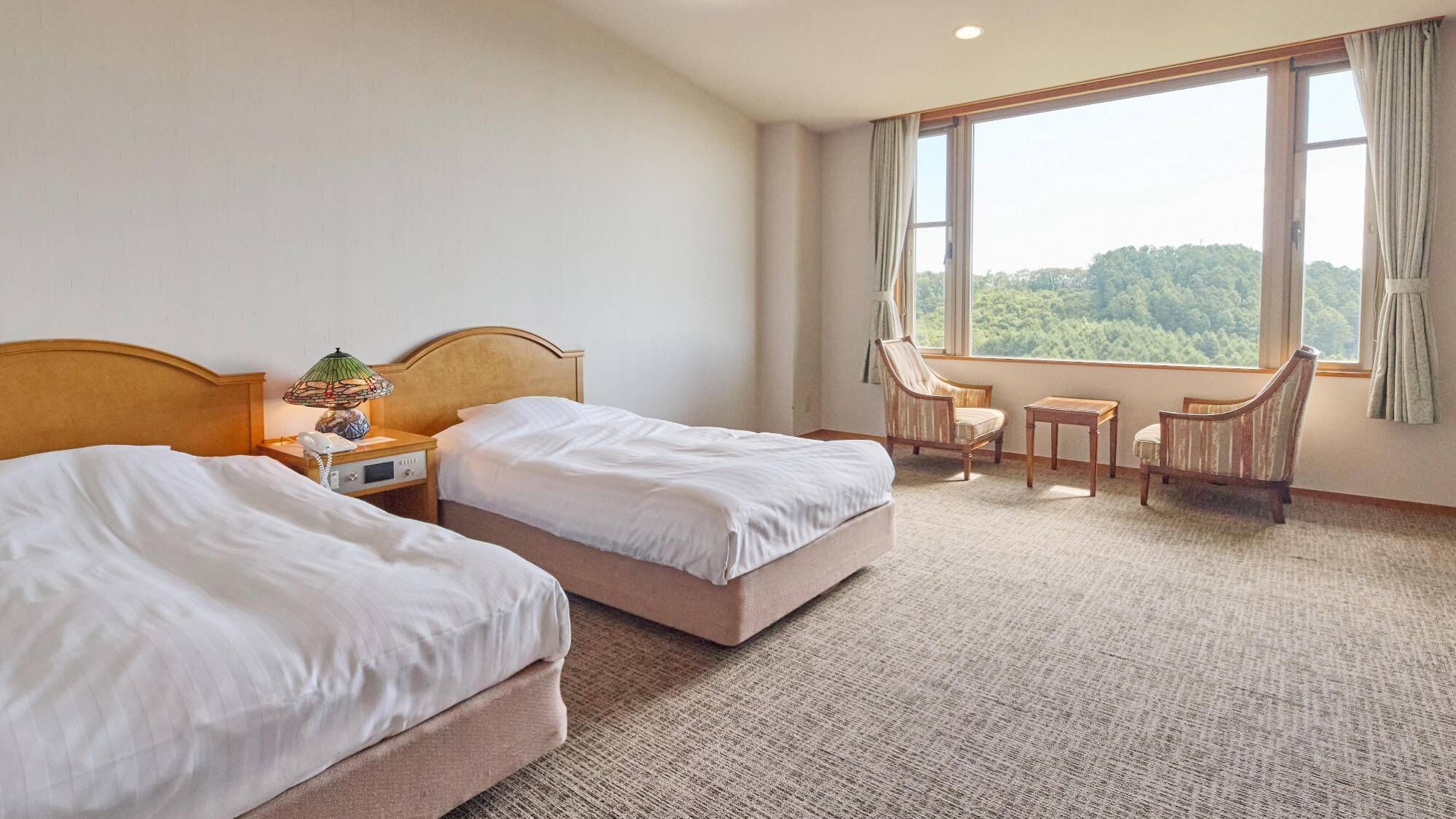 Een hotelkamer met twee bedden, een tafeltje, een stoel en een raam met uitzicht op bomen.
