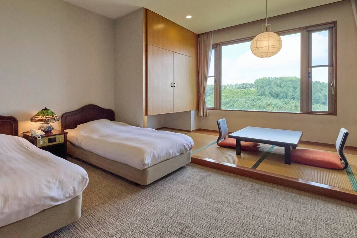 Ein Hotelzimmer mit zwei Betten, einem kleinen Tisch, einem Stuhl, einer Lampe und Blick auf Grün hinter dem Fenster.