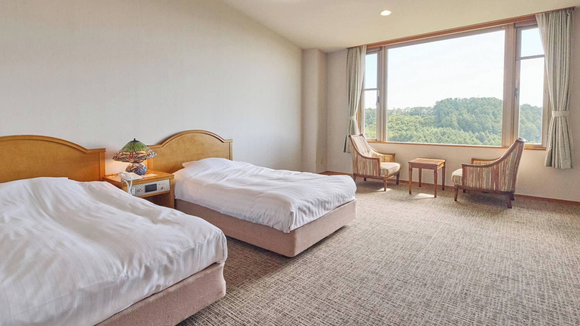 A hotel room with two beds, a small table, a chair, and a window with a view of trees.