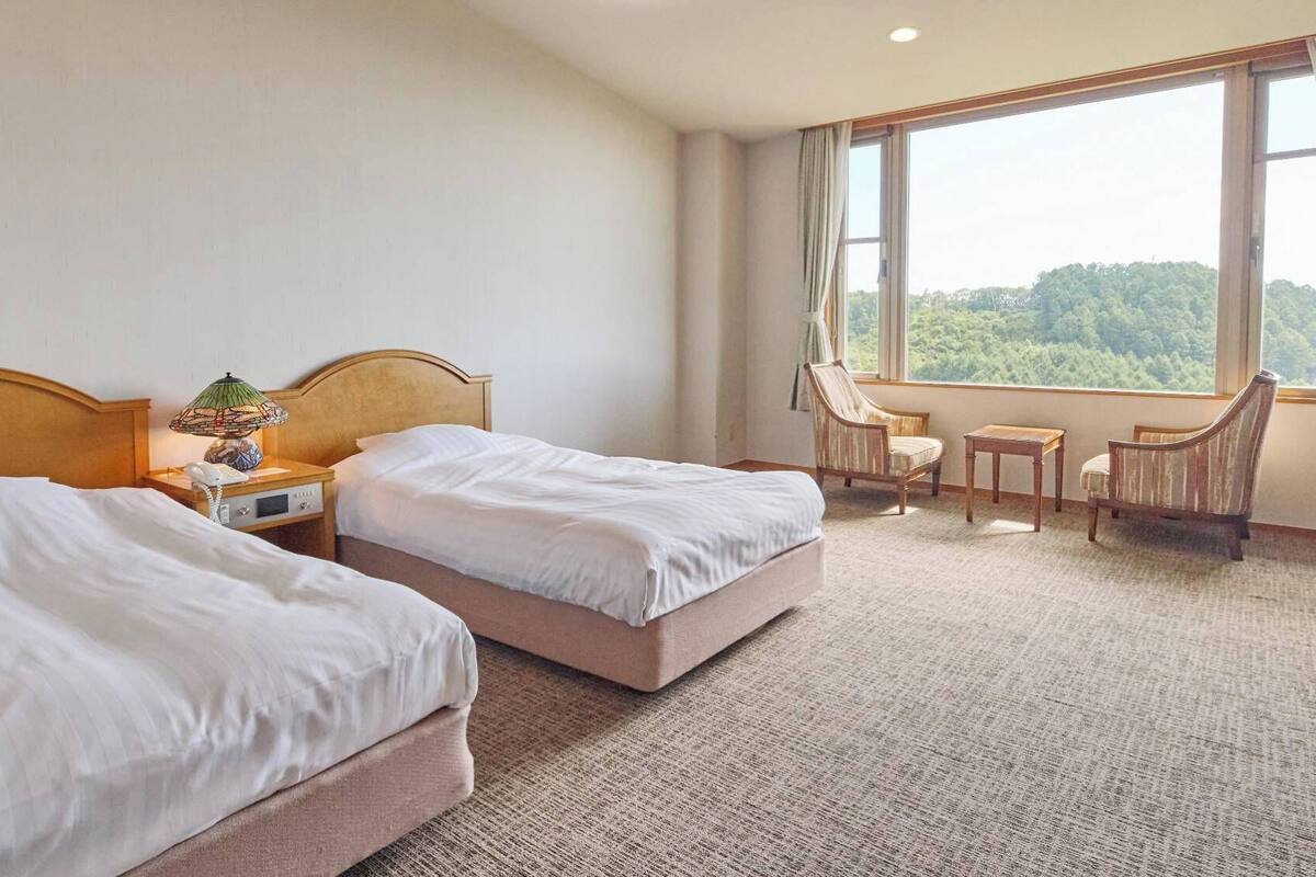 Ein Hotelzimmer mit zwei Betten, einem kleinen Tisch, einem Sessel und einem Fenster mit Blick auf Bäume.