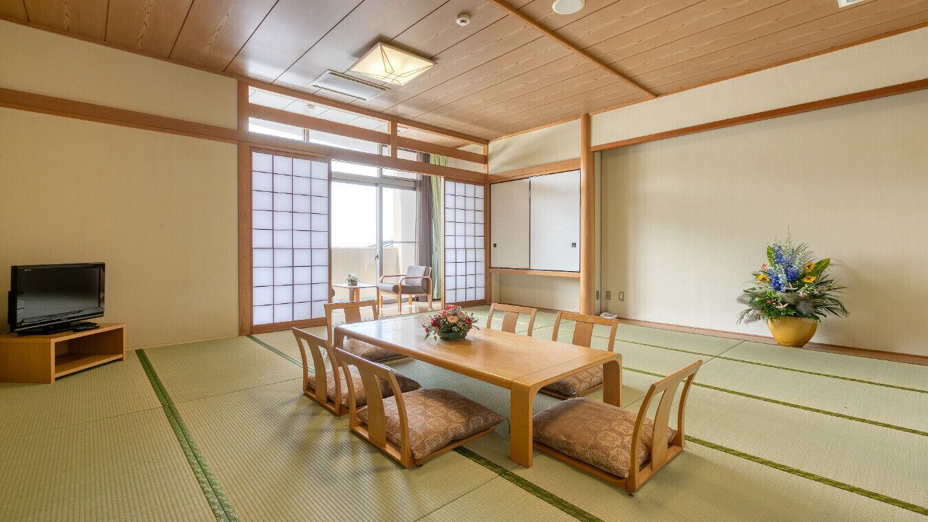 Ein traditioneller japanischer Raum mit Tatami-Bodenbelag, einem niedrigen Tisch mit Kissen, einem Fernseher auf einem Standfuß, Schiebetüren und einer Zimmerpflanze.