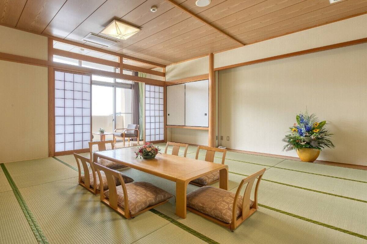 A traditional Japanese room with tatami flooring, a low table with cushions, a television on a stand, sliding doors, and a potted plant.