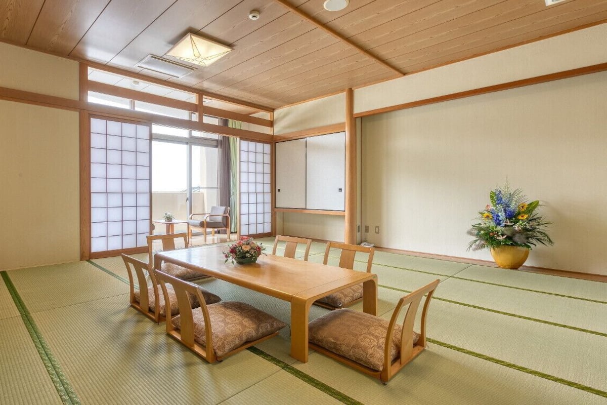 Una habitación tradicional japonesa con suelo de tatami, una mesa baja con cojines, un televisor sobre un mueble, puertas correderas y una planta en maceta.