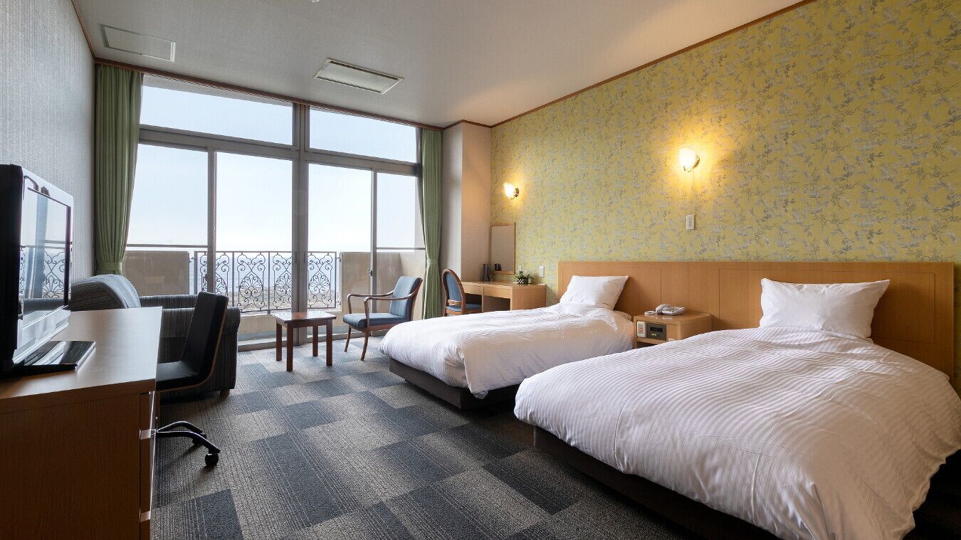Ein Hotelzimmer mit zwei Betten, einem Schreibtisch mit Stuhl, einem kleinen Tisch, einem Fernseher und einem Balkon mit Geländer.