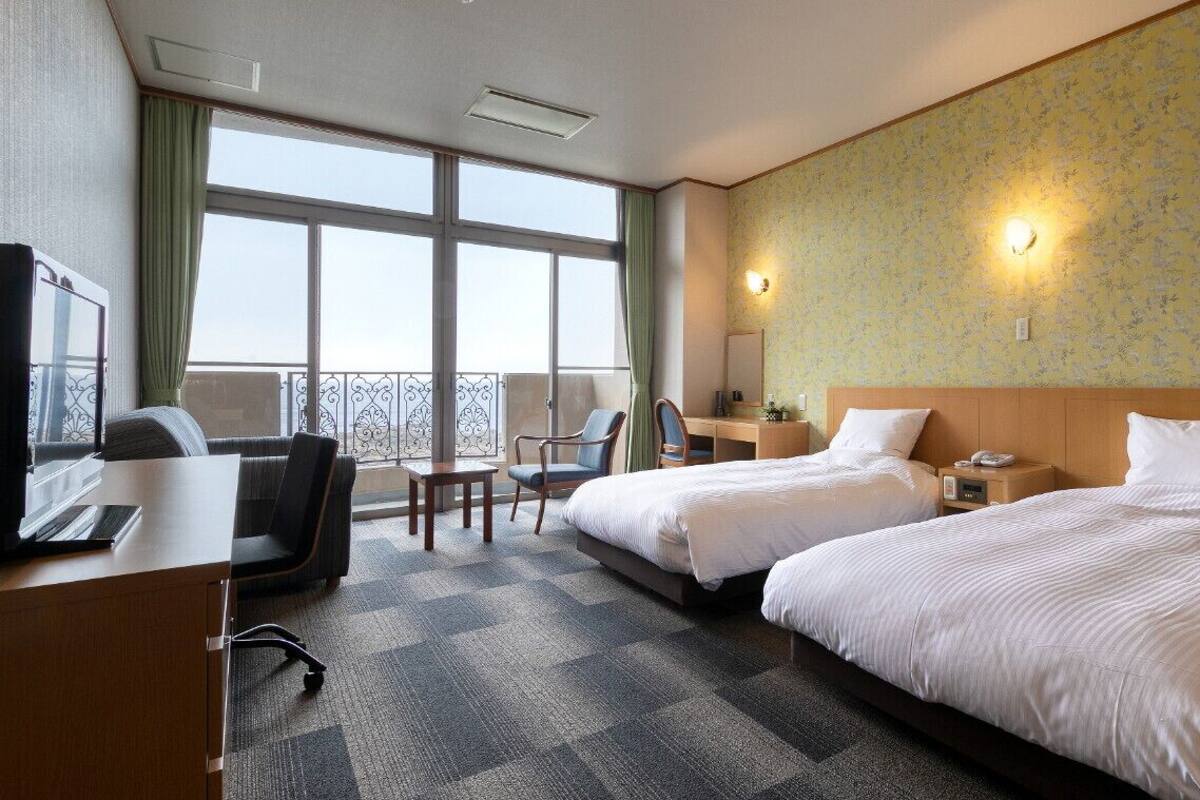 A hotel room with two beds, a desk with a chair, a small table, a TV, and a balcony with a railing.