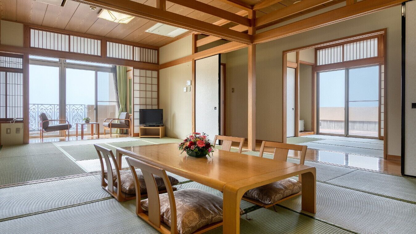 Ein traditioneller japanischer Raum mit Tatami-Böden, einem Holztisch mit Kissen, einem Fernseher und einer Balkontür.