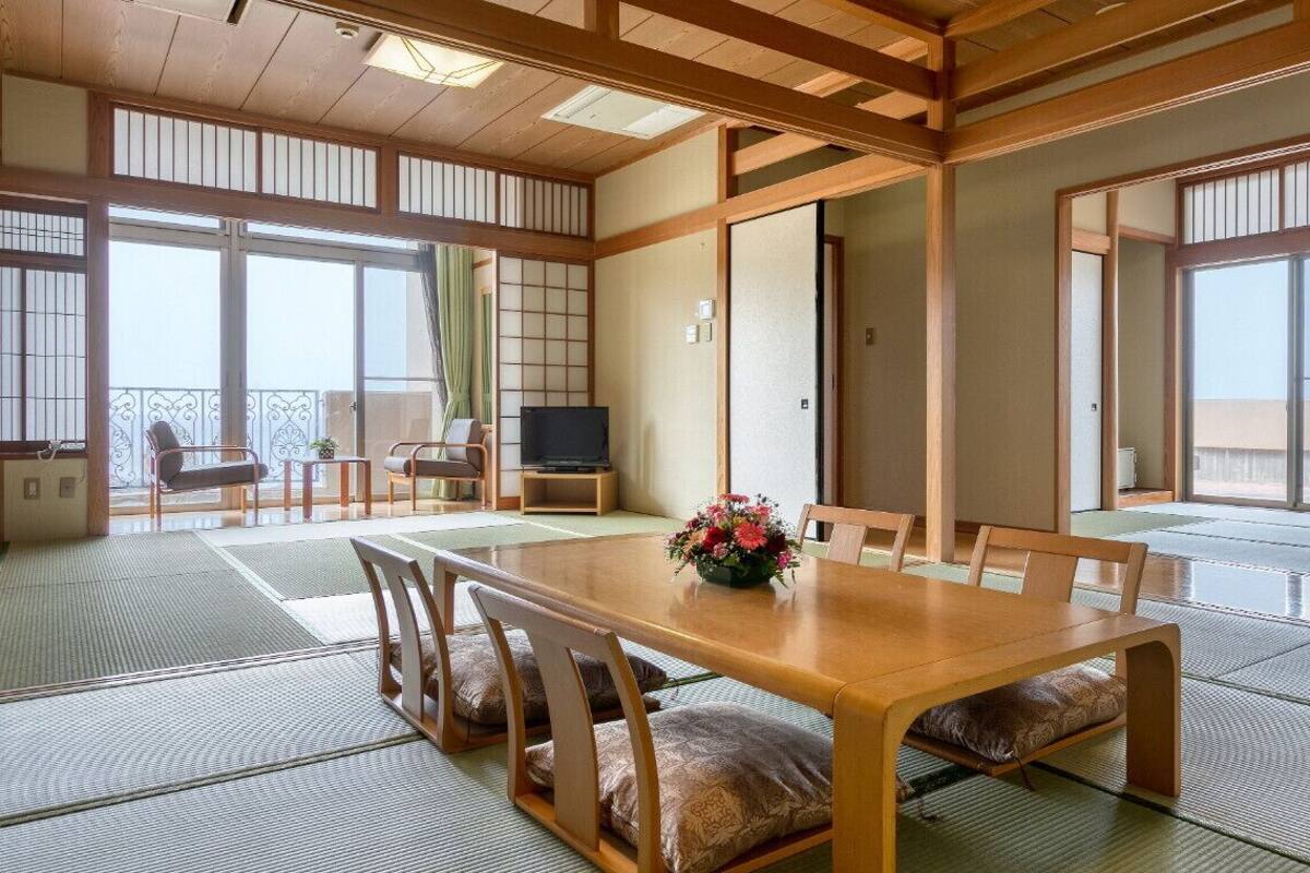A traditional Japanese room with tatami flooring, a wooden dining table with cushions, a television, and sliding doors leading to a balcony.