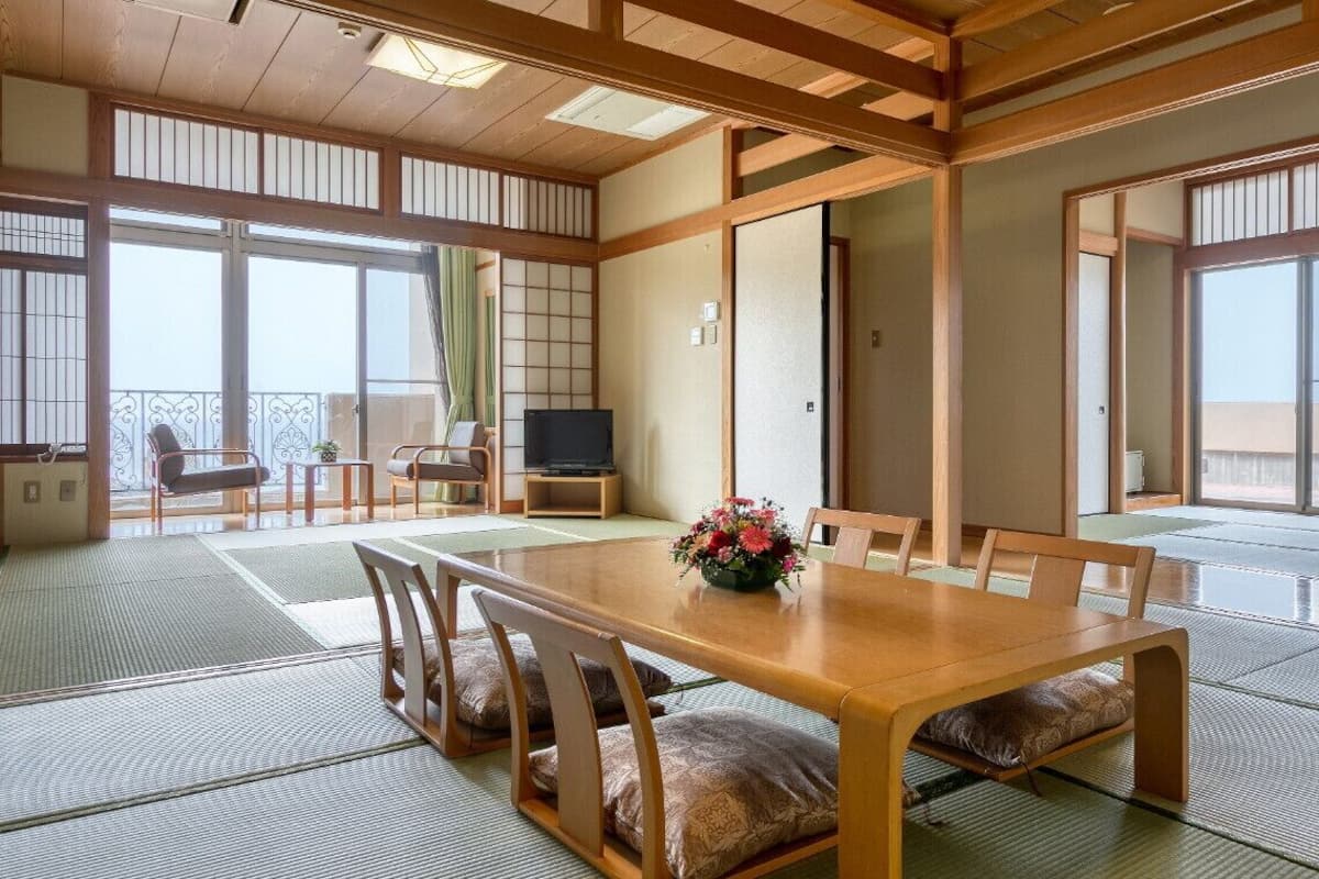 Una habitación japonesa tradicional con suelo de tatami, una mesa de comedor de madera con cojines, un televisor y puertas correderas que dan a un balcón.