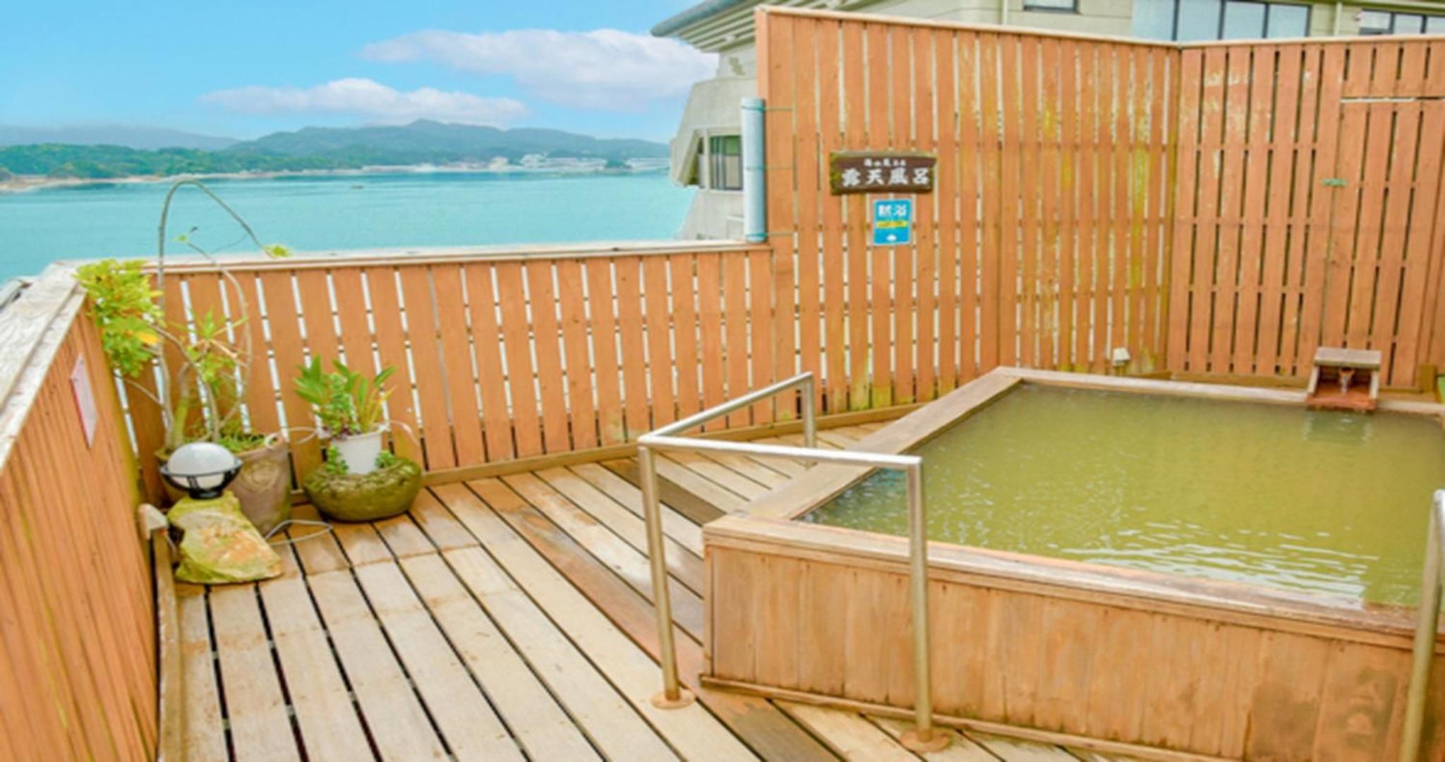 Una terraza de madera con un jacuzzi y plantas en macetas, con vistas a un cuerpo de agua y montañas lejanas.