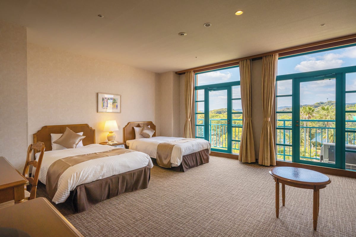 Ein Hotelzimmer mit zwei Betten, einem Schreibtisch, einem Stuhl, einem kleinen Tisch und großen Fenstern mit Blick auf Grünflächen.