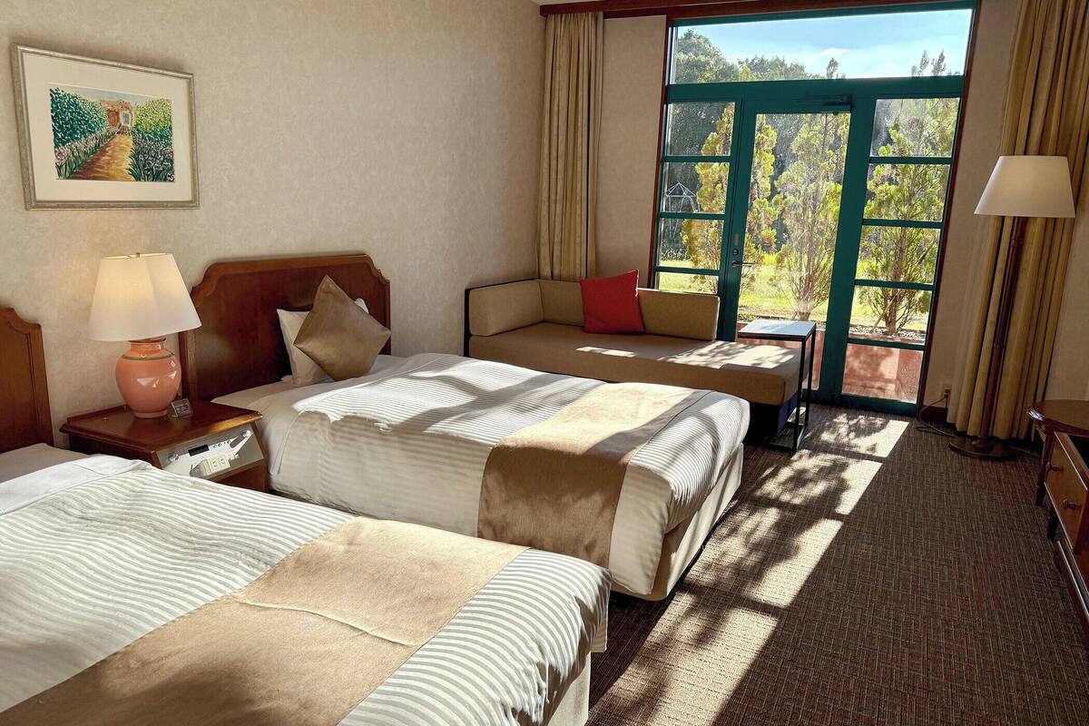 Une chambre d’hôtel avec deux lits, une table de chevet, une lampe, une fenêtre avec des rideaux et une vue sur les arbres.