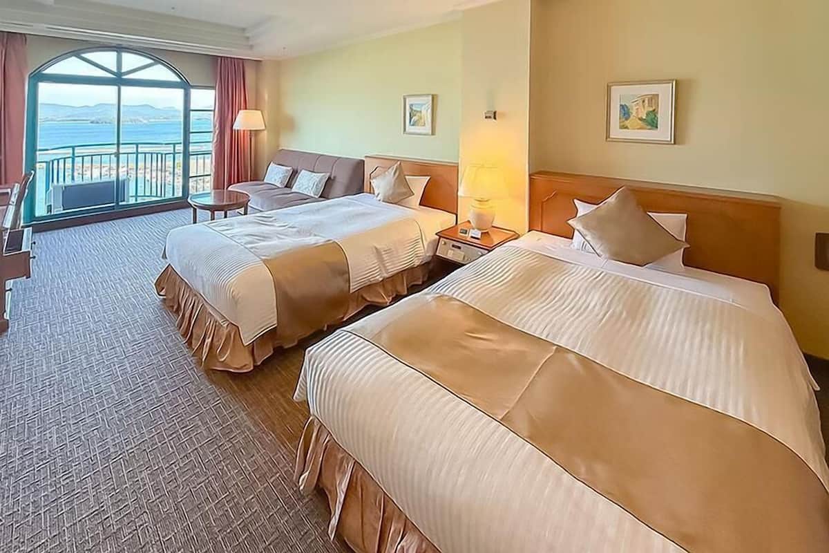 Ein Hotelzimmer mit zwei Betten, einem Schreibtisch, einem Sessel und Blick auf das Meer.